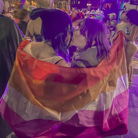 Max and Chloe kiss while holding a lesbian flag that is draped around them. They are at a pride parade.