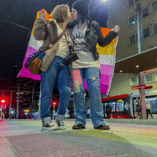 Max and Chloe kiss while holding a lesbian flag that is draped around them. They stand on a footpath that is painted rainbow.