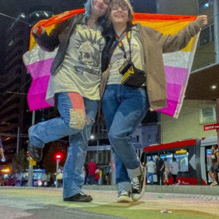Max and Chloe kiss while holding a lesbian flag that is draped around them. They stand on a footpath that is painted rainbow.