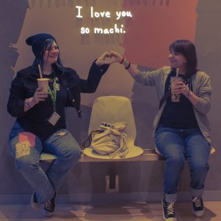 Max and Chloe holding their hands out to make a heart together, underneath a sign that says "I love you so machi". Max and Chloe are wearing matching shirts that says "Partner in Crime" (with a skull), and "Partner in Time" (with a butterfly). They are both holding their own bubble tea.