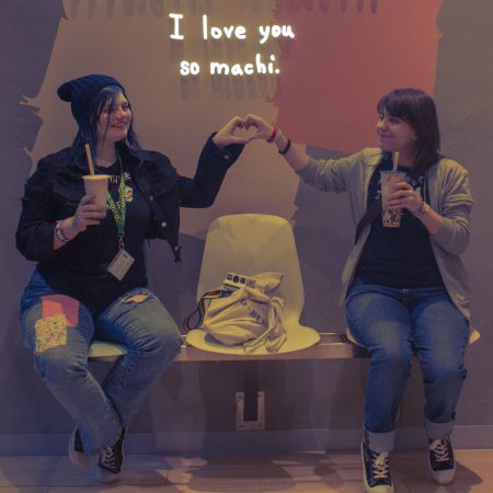 Max and Chloe holding their hands out to make a heart together, underneath a sign that says "I love you so machi". Max and Chloe are wearing matching shirts that says "Partner in Crime" (with a skull), and "Partner in Time" (with a butterfly). They are both holding their own bubble tea.