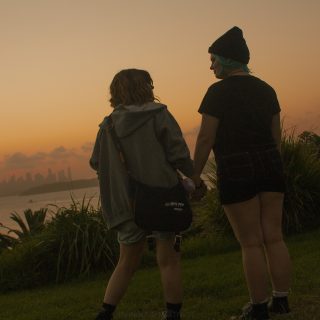 Max and Chloe hold hands and look at each other, while facing the ocean, and views of the city can be faintly seen beyond them. Their backs are toward the camera. The sky is golden and sun is setting.